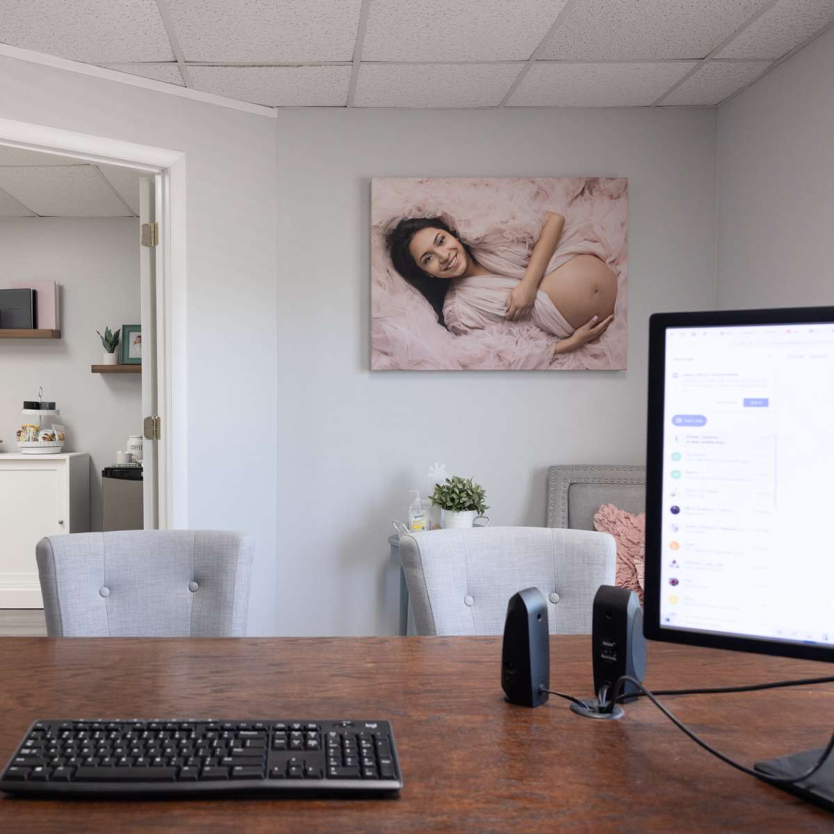 A modern office space with a wooden desk, a computer monitor, and two light-colored chairs, leading to an adjoining room with a sink and shelf.