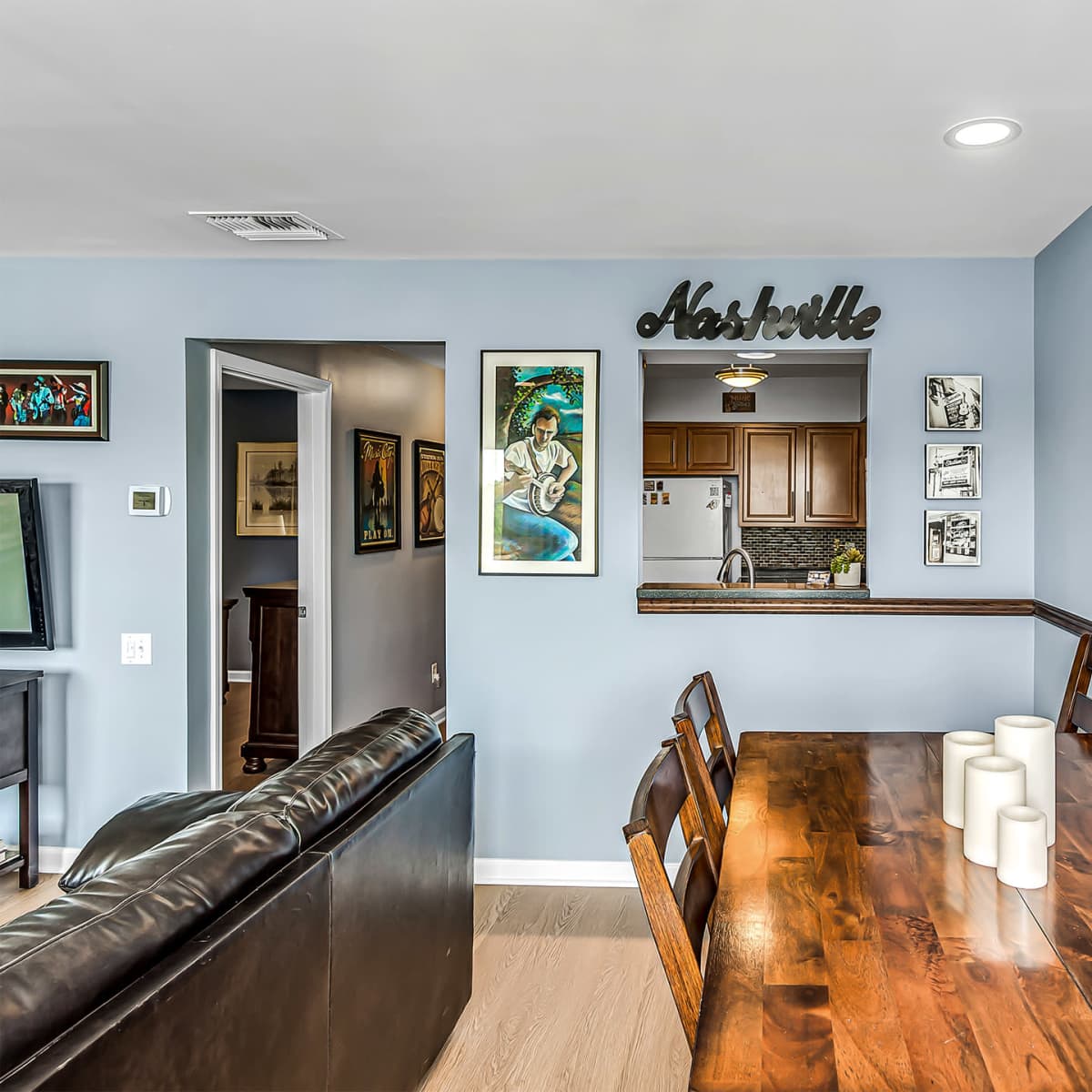A dining area featuring a wooden table with candles, framed artwork on the walls, and a view into a kitchen area.