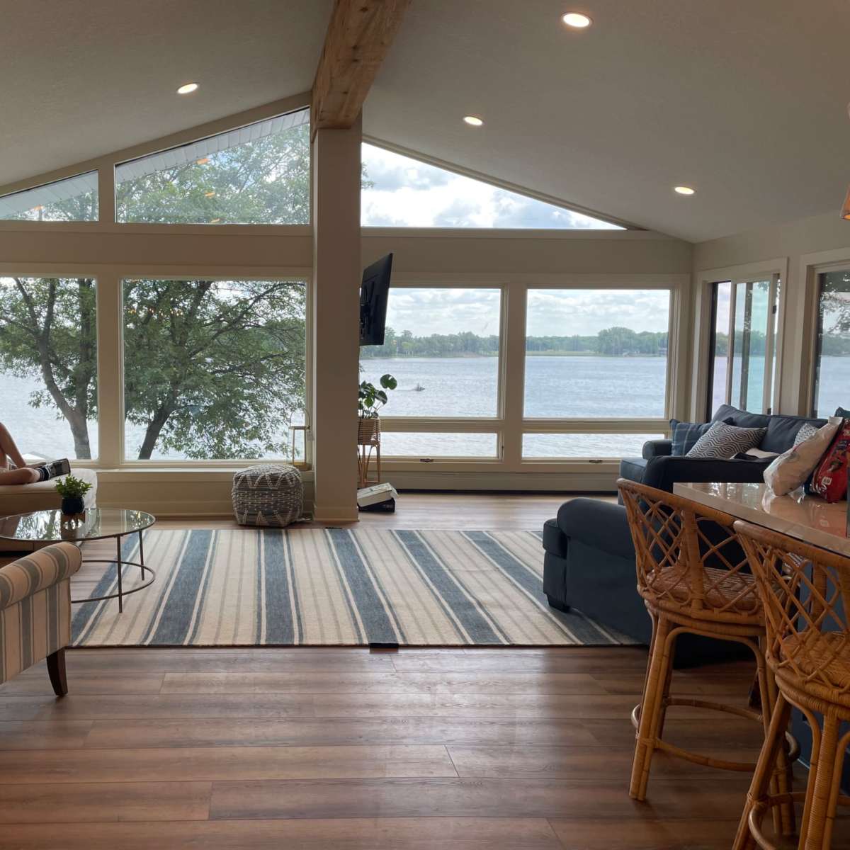 A modern living room features large windows overlooking a lake, with a comfortable seating area, a coffee table, and a striped rug.