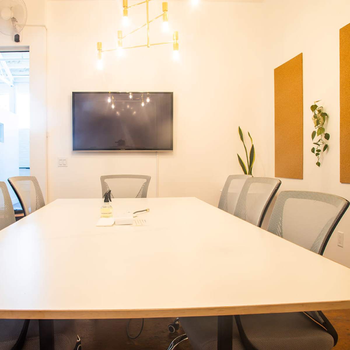 A conference room features a long white table surrounded by mesh chairs, with a TV mounted on the wall and plants in the corners.
