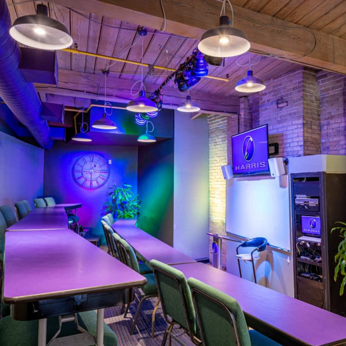 A modern conference room features long purple tables, green chairs, and a wall-mounted screen with the "HARRIS" logo, illuminated by hanging lights.
