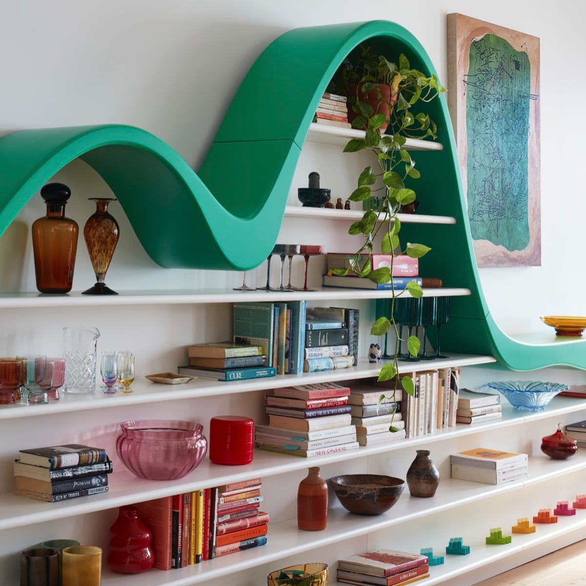 A wavy green bookshelf displays various books, decorative bottles, and plants against a light-colored wall.