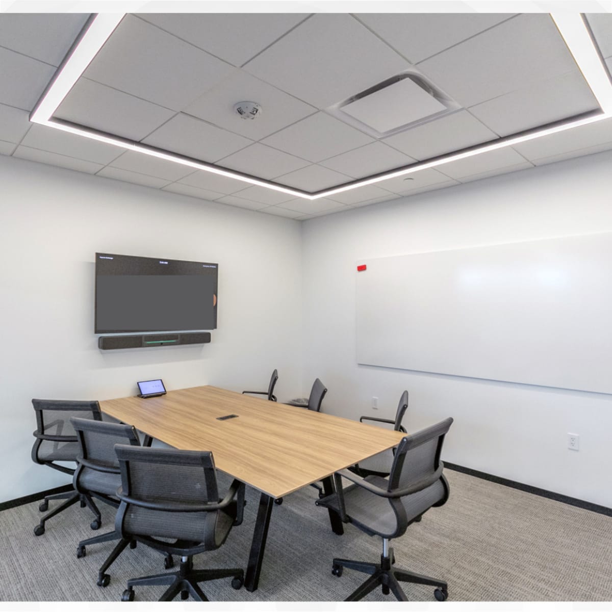 A meeting room featuring a rectangular wooden table surrounded by six office chairs, with a flat-screen TV on the wall and a whiteboard nearby.
