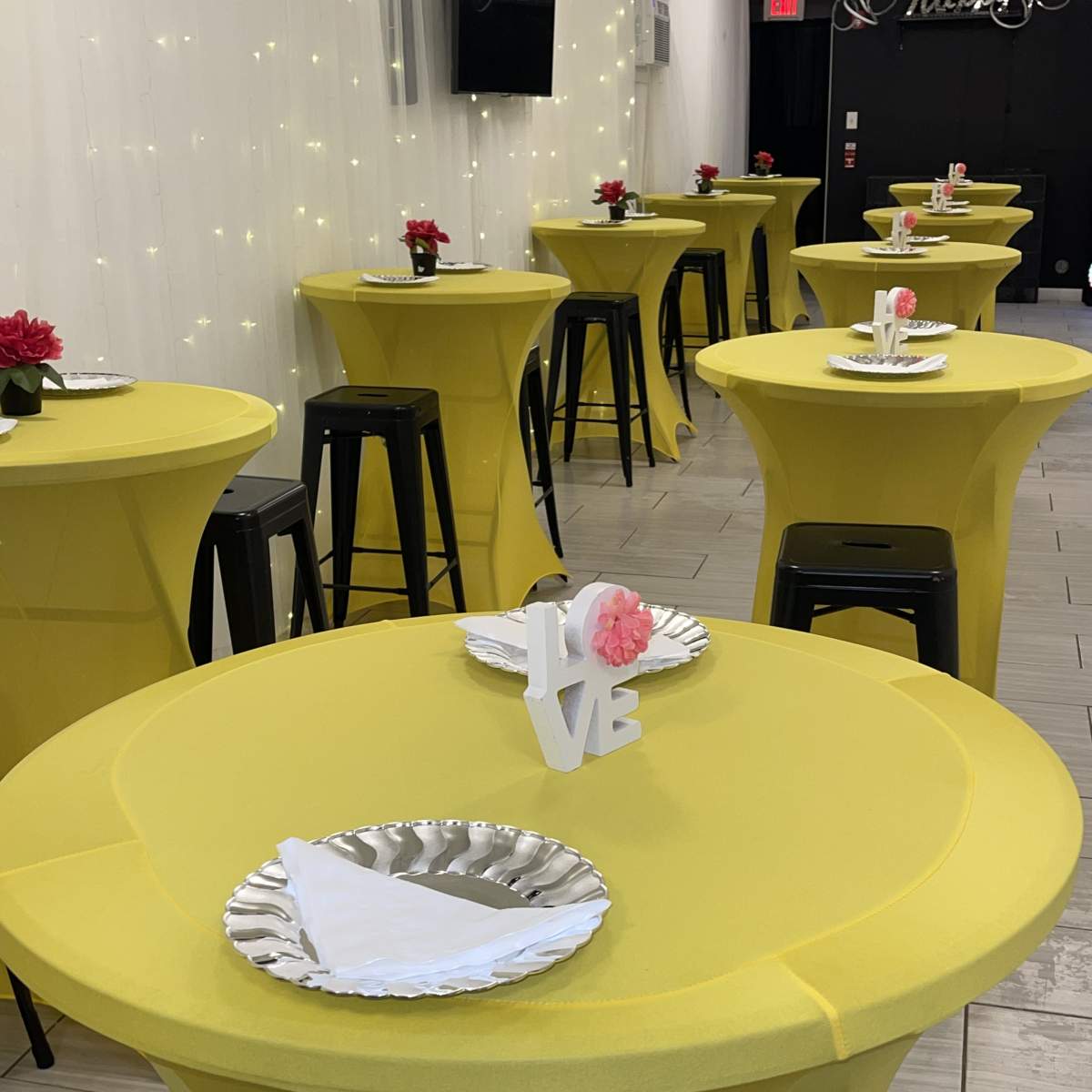 A room with several round yellow tables, each set with silver plates, napkins, and decorative flowers.