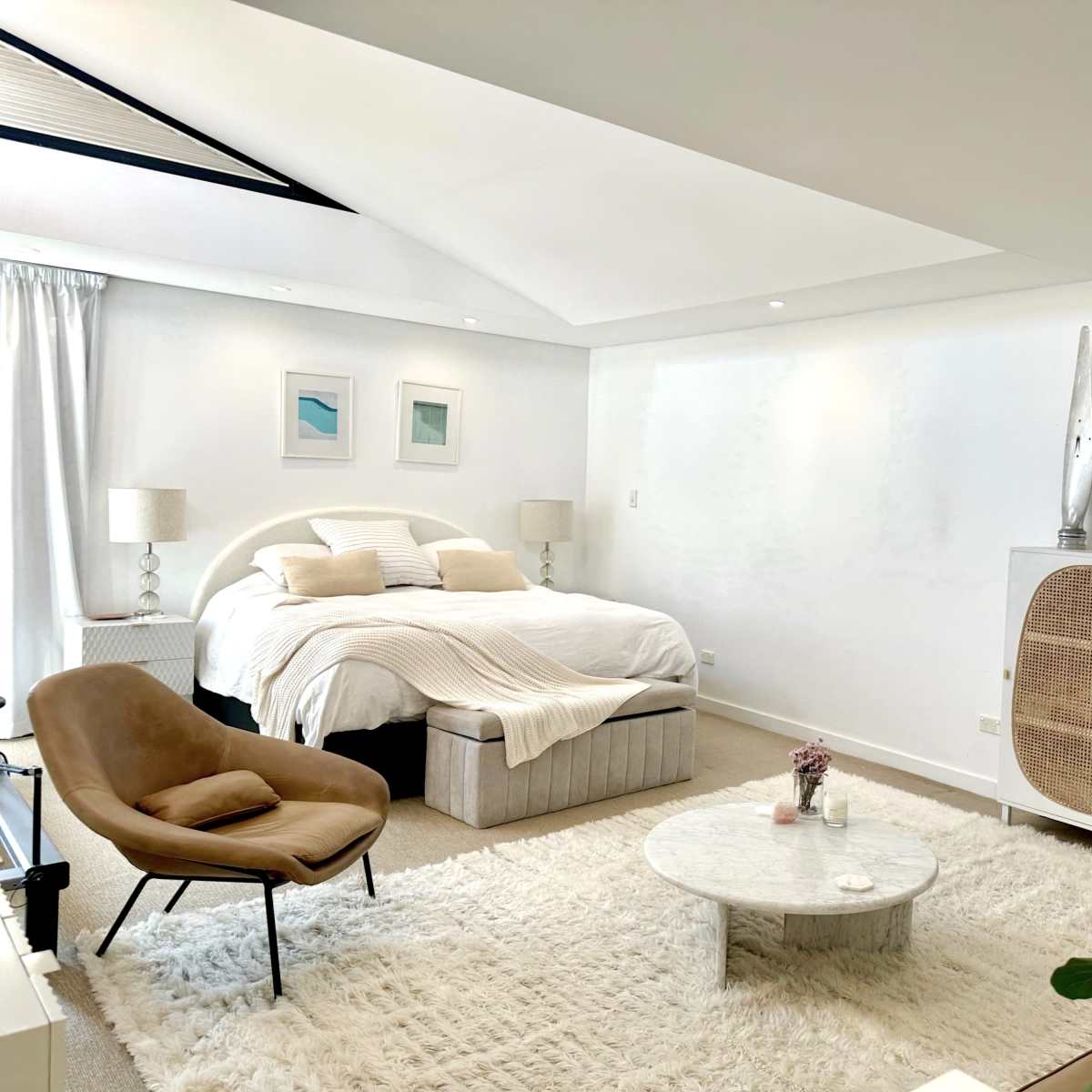 A modern bedroom features a large bed with striped bedding, a circular coffee table on a plush rug, and art displayed on the walls.