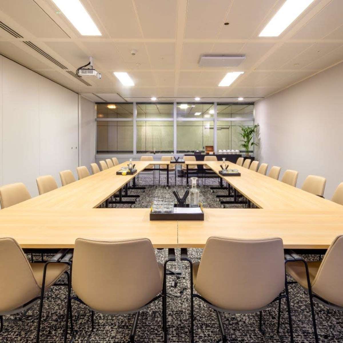 A large, rectangular conference room features a long wooden table surrounded by beige chairs, with a projector and glass windows in the background.