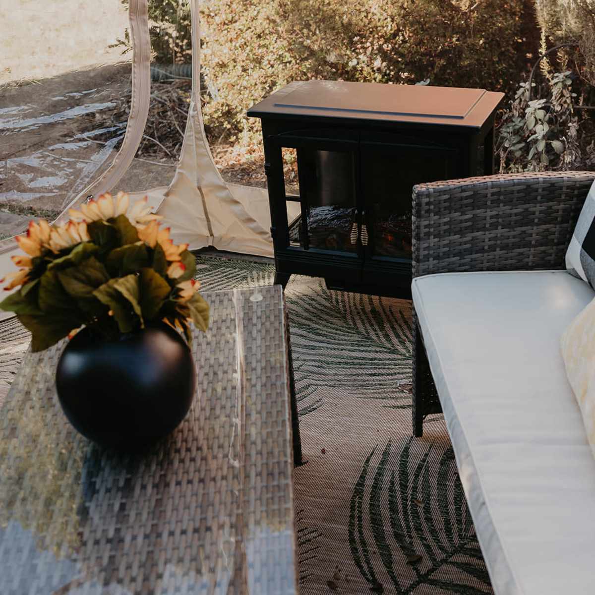 A cozy outdoor seating area features a wicker sofa with cushions, a decorative table, and a portable heater, all set within a transparent tent.