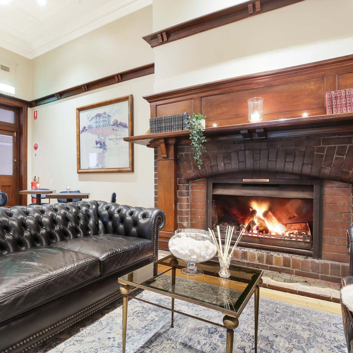 A cozy lounge area featuring a leather chesterfield sofa, a glass coffee table, and a brick fireplace with flames.