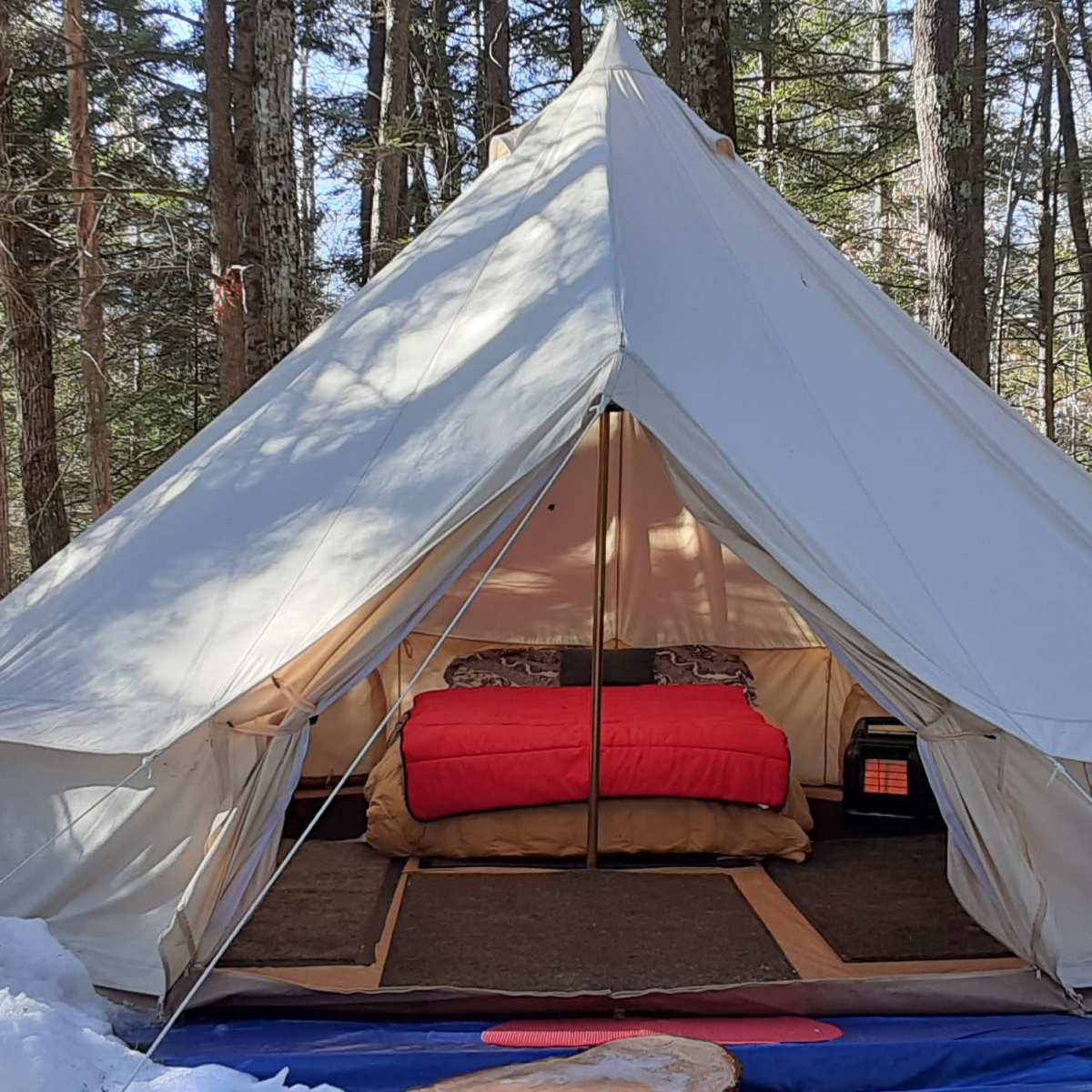 A large white tent is set up in a snowy forest, with a red bed inside and a propane tank outside.