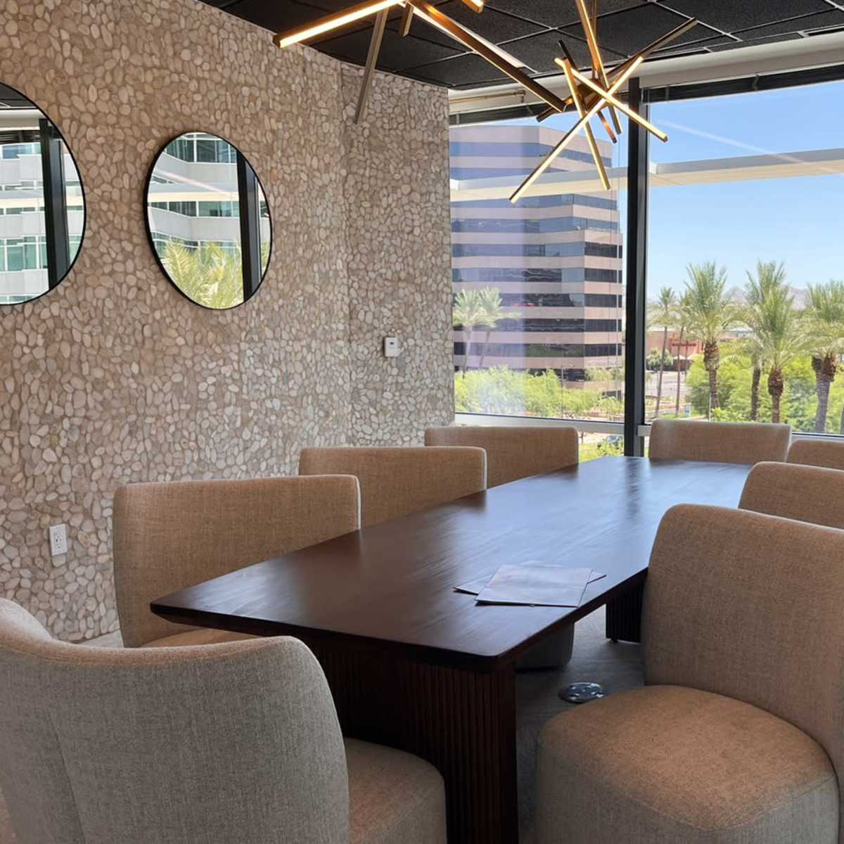 A modern conference room features a large wooden table surrounded by upholstered chairs, with large windows overlooking palm trees.