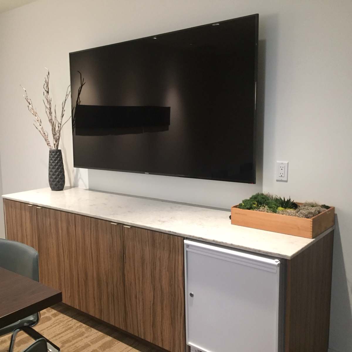 A wall-mounted television is displayed above a wooden cabinet that features a small refrigerator and a planter with greenery.