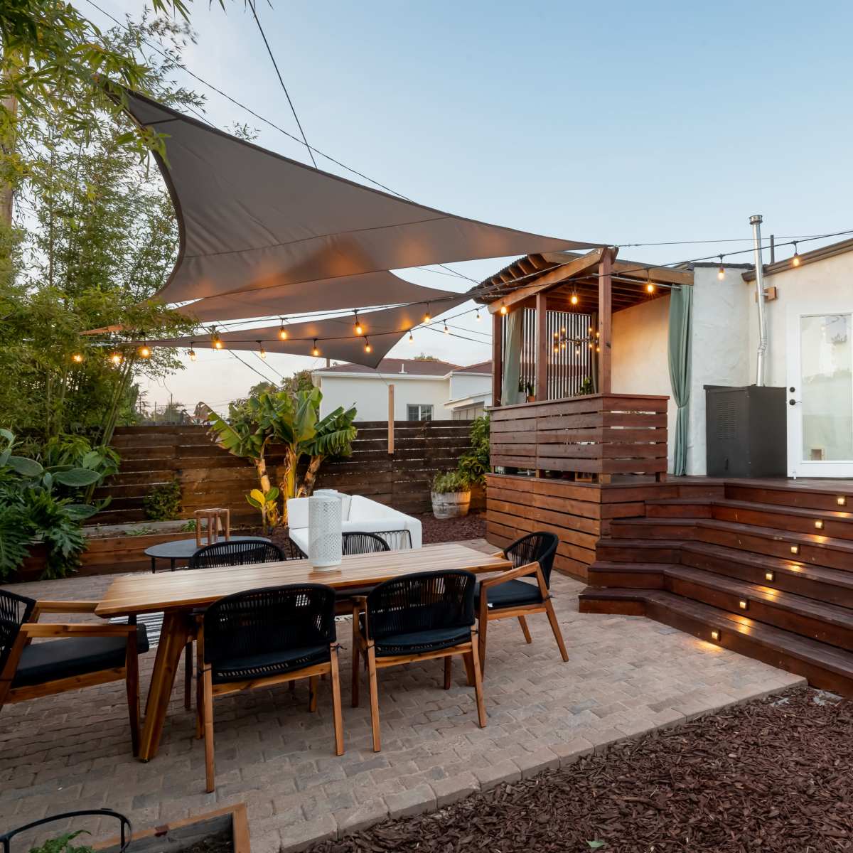 A patio area features a wooden dining table with black chairs, surrounded by plants, leading up to a house with an open door and a shade sail above.