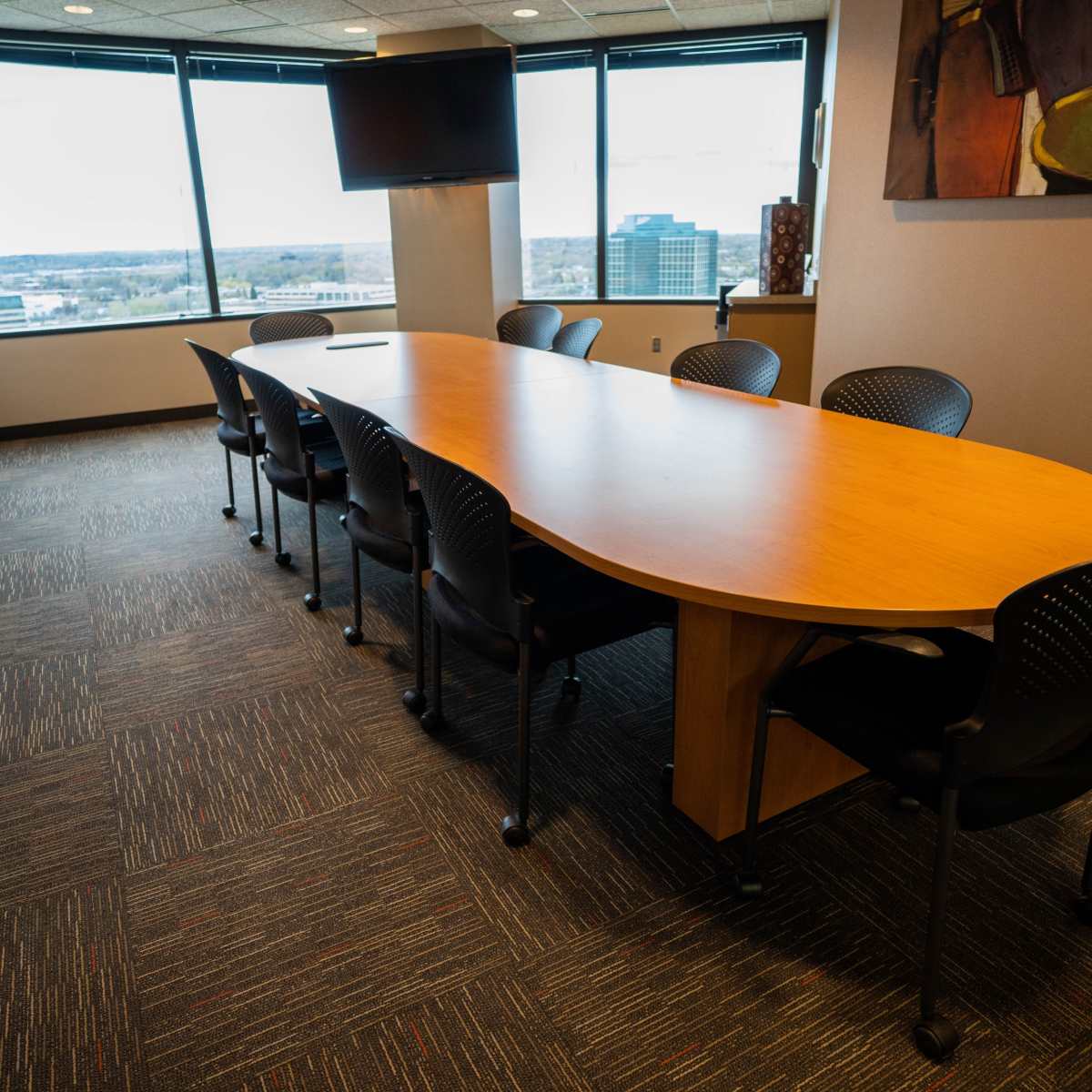 A long, oval conference table with black chairs is positioned in a modern meeting room surrounded by large windows.
