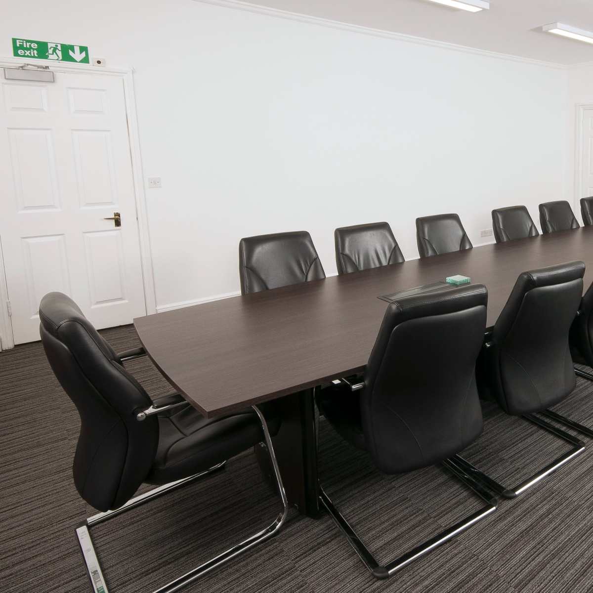 A large conference room features a long wooden table surrounded by black leather chairs, with a wall-mounted television and exit door visible.