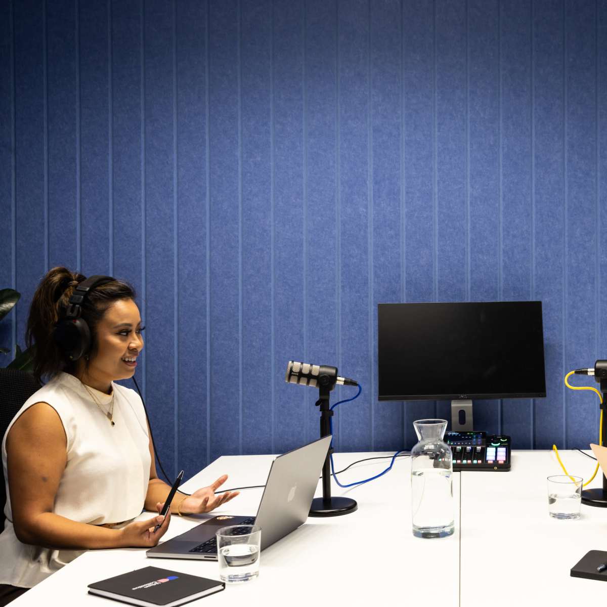 Two women are seated at a table with laptops and microphones, engaged in conversation in a blue-walled podcast recording studio.