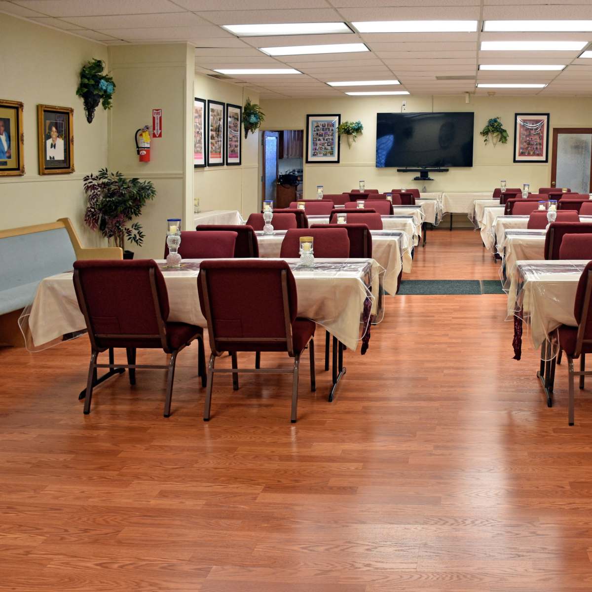 A large dining area features rows of tables covered with light-colored tablecloths and chairs arranged around them, while walls are adorned with framed photographs.