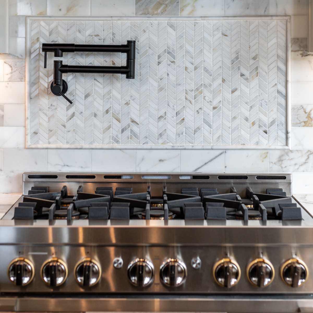 A modern kitchen backsplash with a herringbone pattern in marble above a stainless steel gas stove and a metallic range hood.