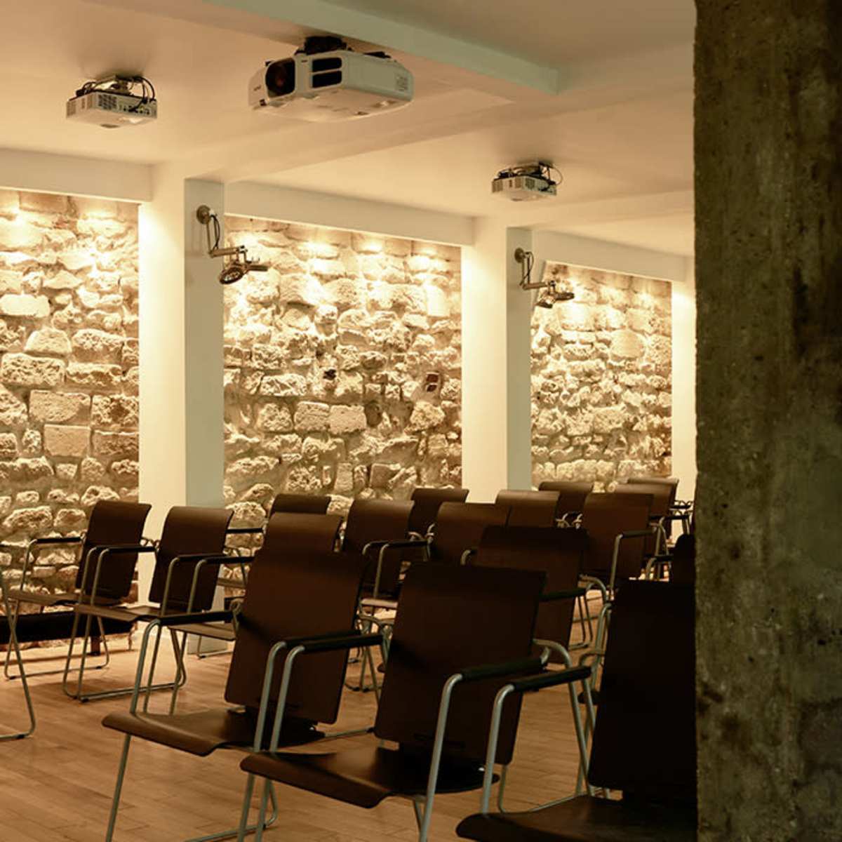 A modern interior space featuring stone walls, wooden floors, and a row of metal chairs facing a projection area, with large windows along one side.