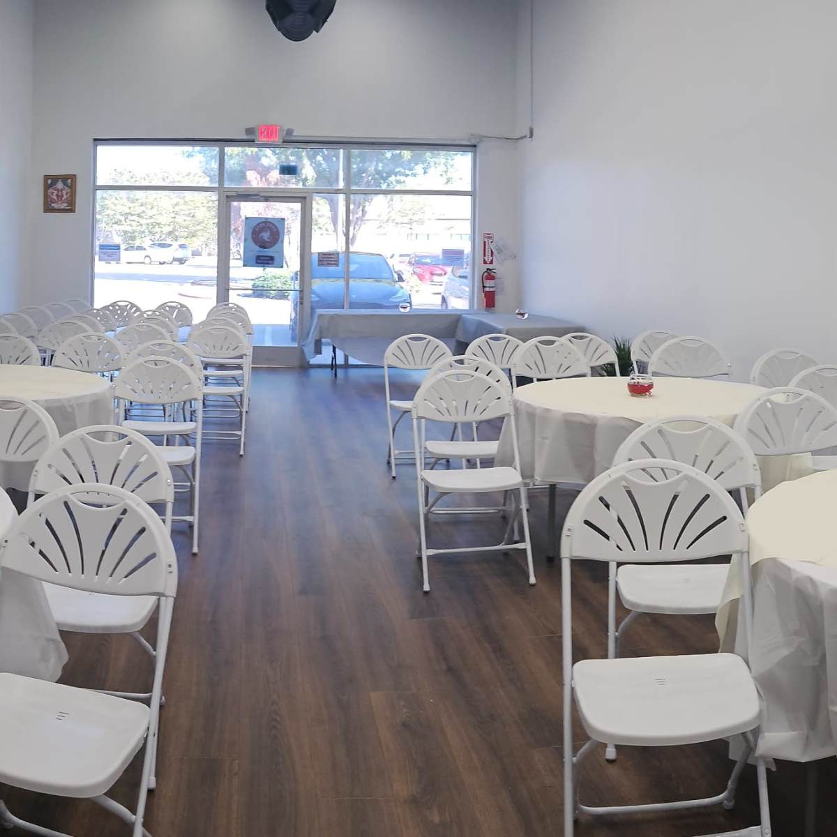 A spacious room arranged with several round tables covered in white tablecloths and surrounded by white chairs, leading to a large front window.