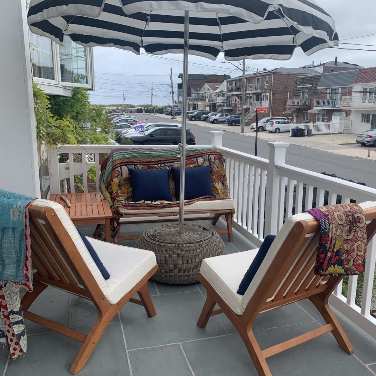 A small porch features two wooden chairs, a round table with an umbrella, and a sofa draped with colorful throws, overlooking a street with nearby houses.