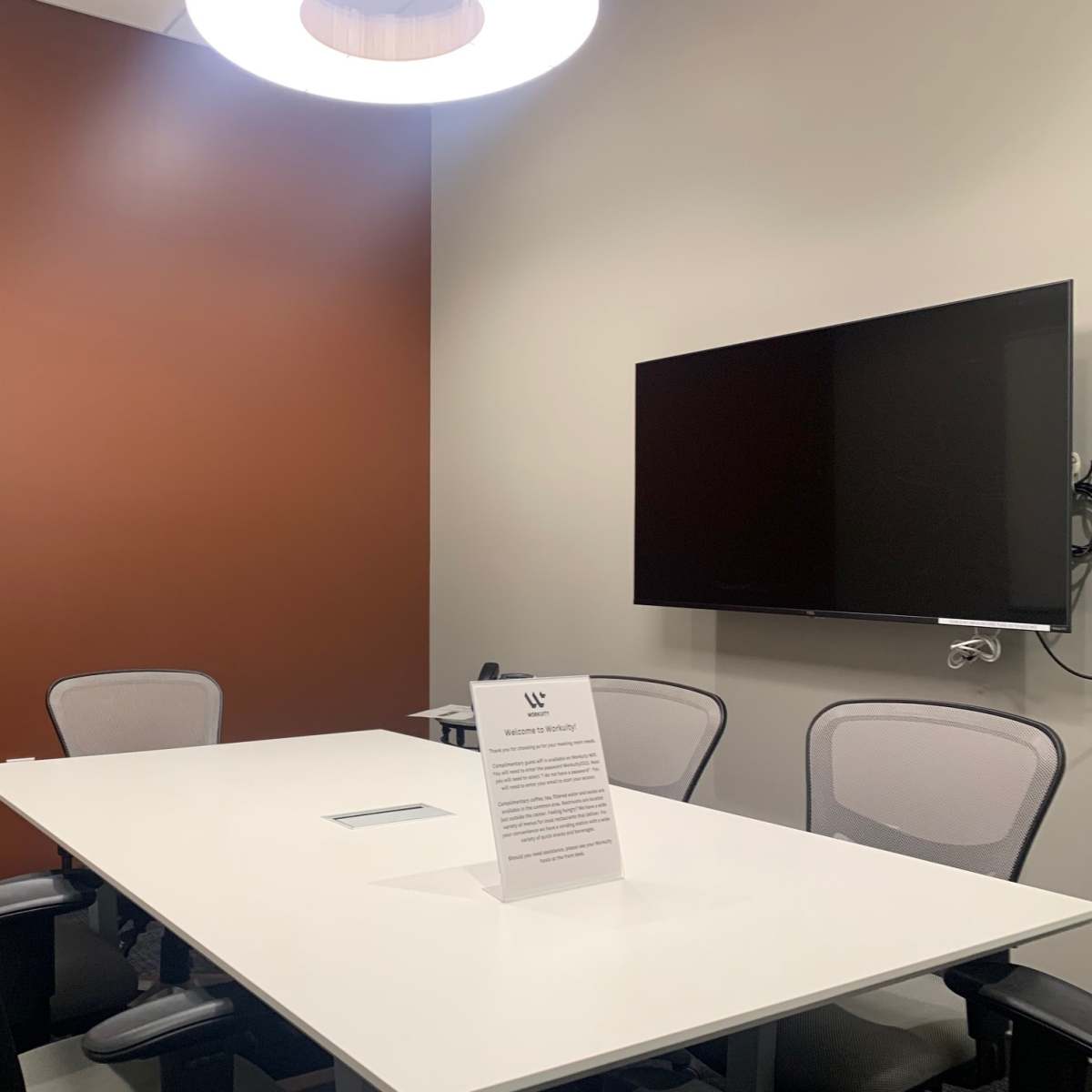 A small meeting room features a rectangular table with four chairs, a wall-mounted TV, and a sign on the table.