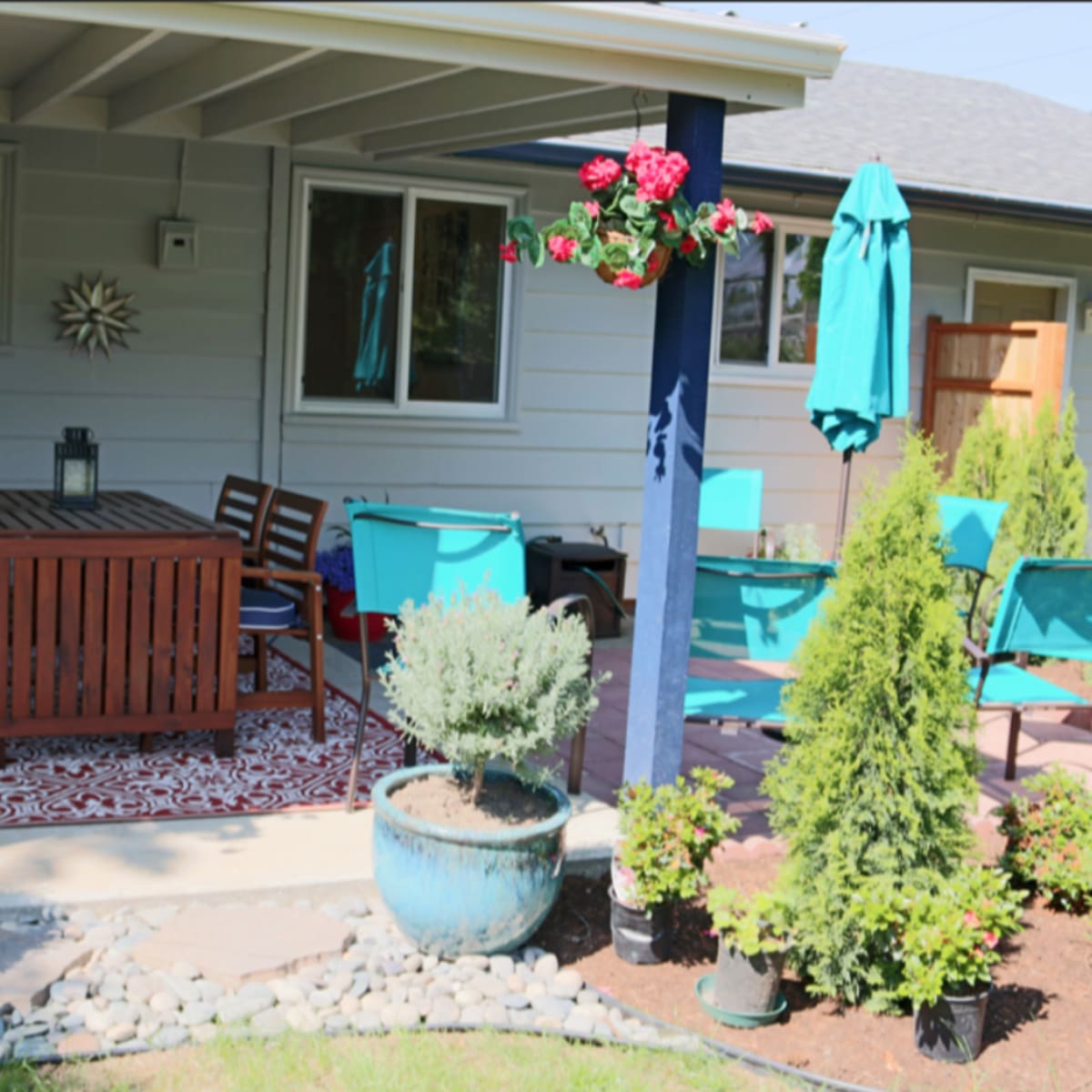 A covered patio area features a wooden dining table with chairs, potted plants, and decorative hanging flowers.