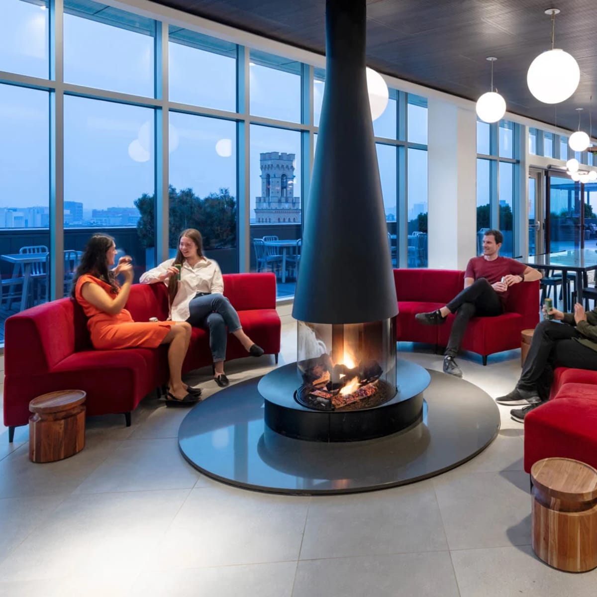 A modern lounge area with comfortable seating around a central fireplace, large windows providing city views, and a few people engaged in conversation.