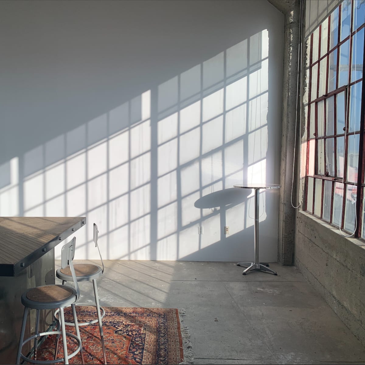 A sunlit room with a wooden bar, two stools, an arcade machine in the corner, and a small table near a large window casting shadows on the wall.