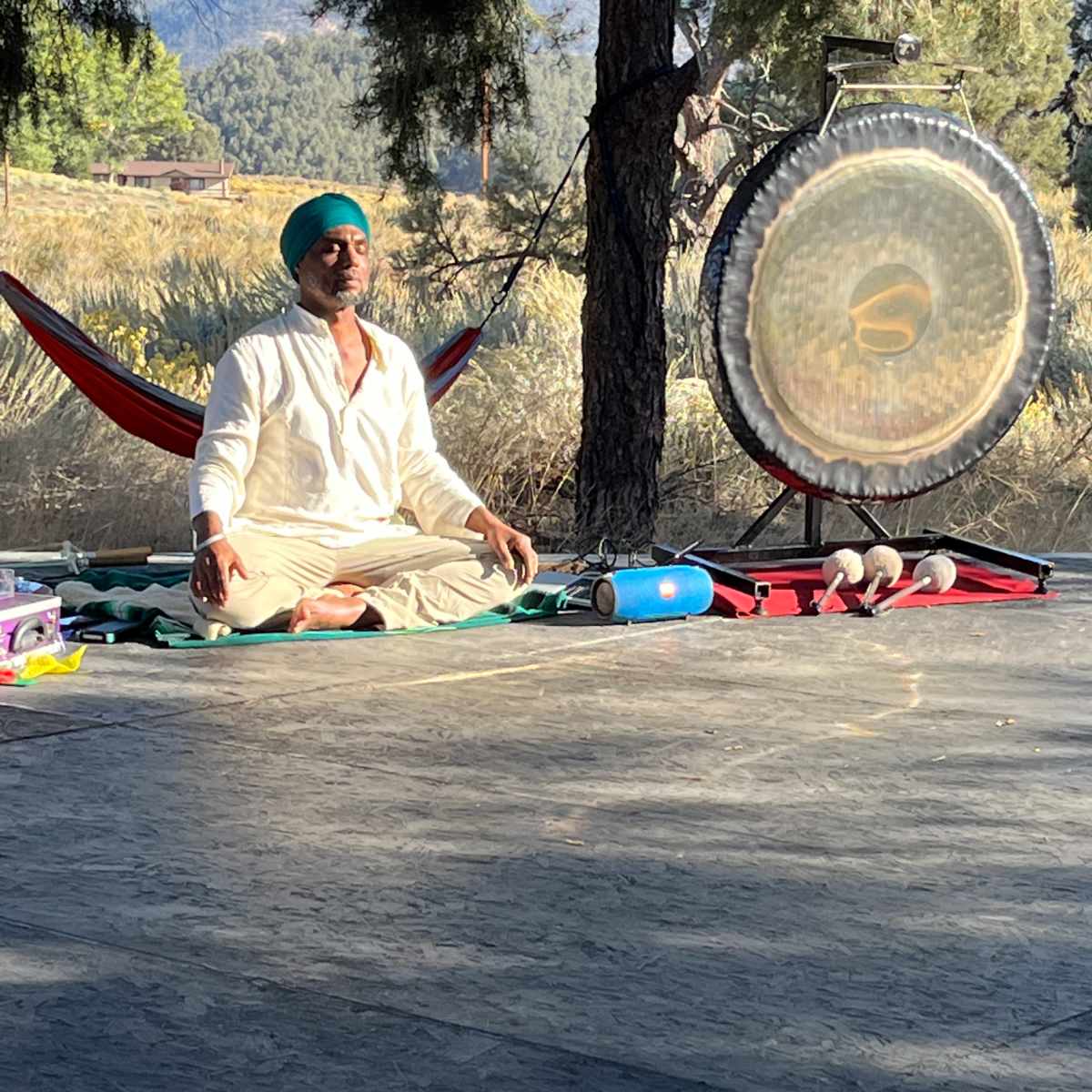 A person sits cross-legged on a wooden platform near a large gong, surrounded by trees and a hammock in the background.