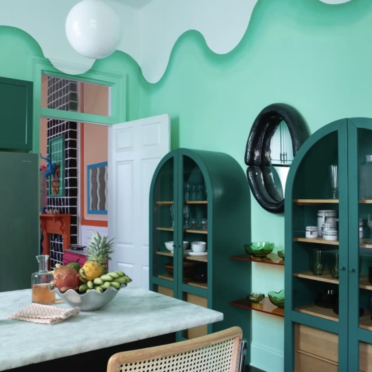 The kitchen features green wave-patterned walls, a marble table, and wooden cabinets displaying various dishware and fresh fruits.