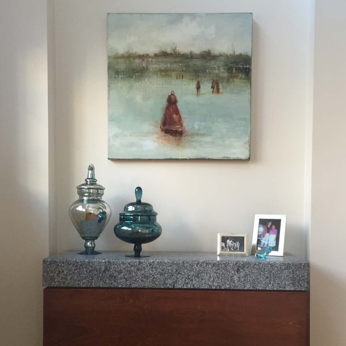 A wooden sideboard with a granite top displays two decorative glass jars, a framed photo, and an abstract painting of figures in a landscape.