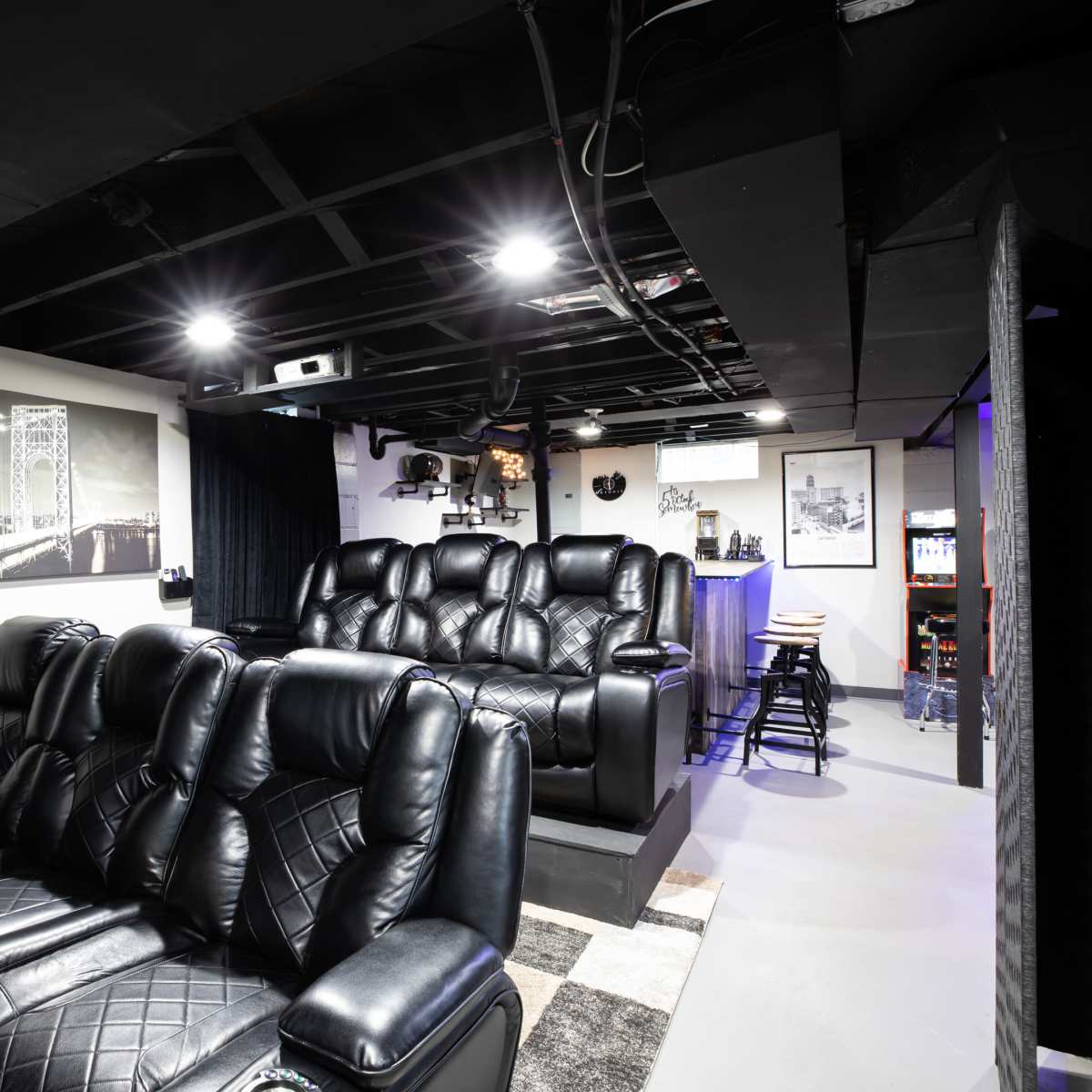 A home theater room with black leather recliners arranged in front of a projection screen, and a snack bar area in the background.