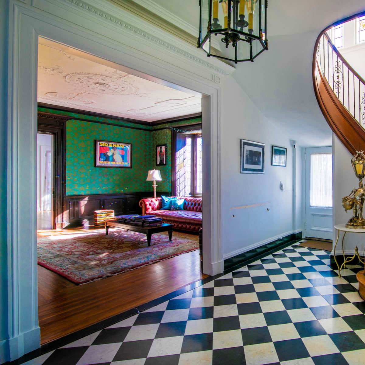 An entryway with a black and white tiled floor leading to a wooden staircase, with a glimpse of a richly decorated living room visible through an open door.