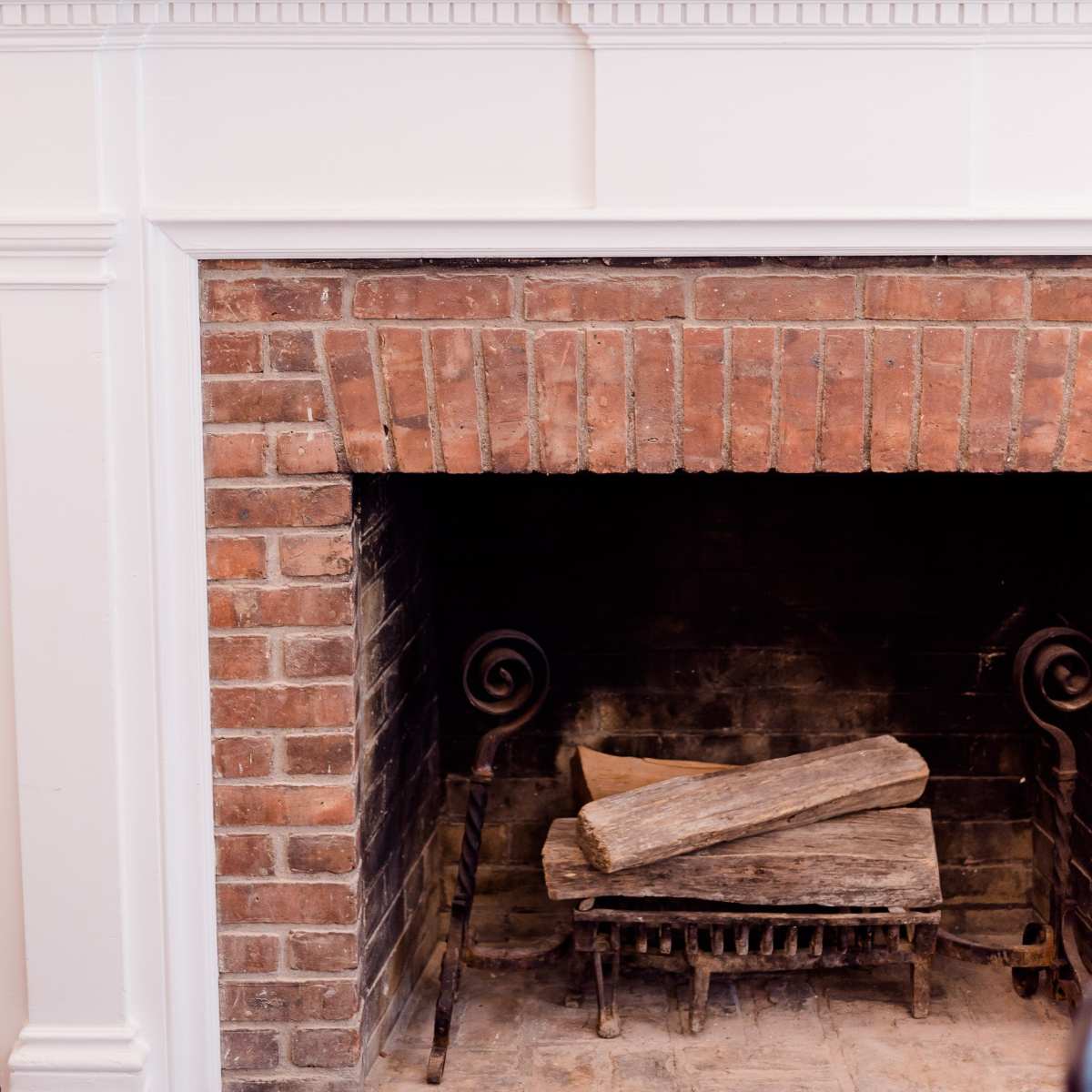 A brick fireplace with a wood log holder contains stacked logs, topped by a white mantel displaying three vases of flowers.