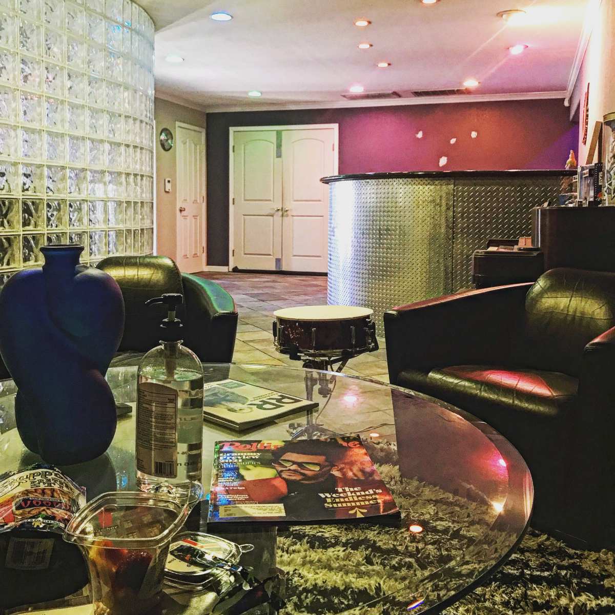 The scene shows a modern interior featuring a glass block wall, a circular bar area, and a coffee table with drinks and magazines in a lounge setting.