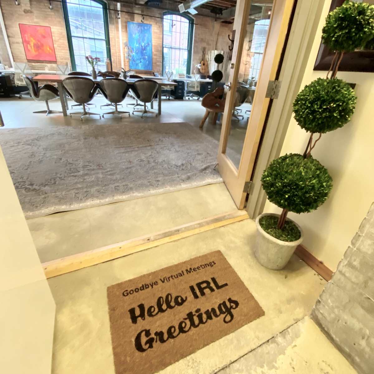 A doorway with a welcome mat stating "Hello IRL Greetings," leading into a spacious room with modern furniture and artwork on the walls.