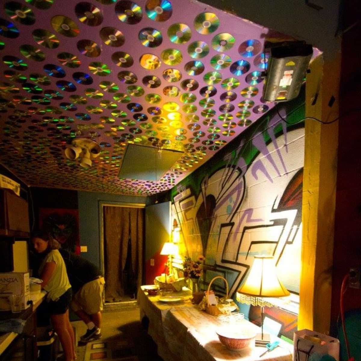 A small kitchen area features a ceiling adorned with colorful CDs, a table set with various items, and a person preparing food at the counter.