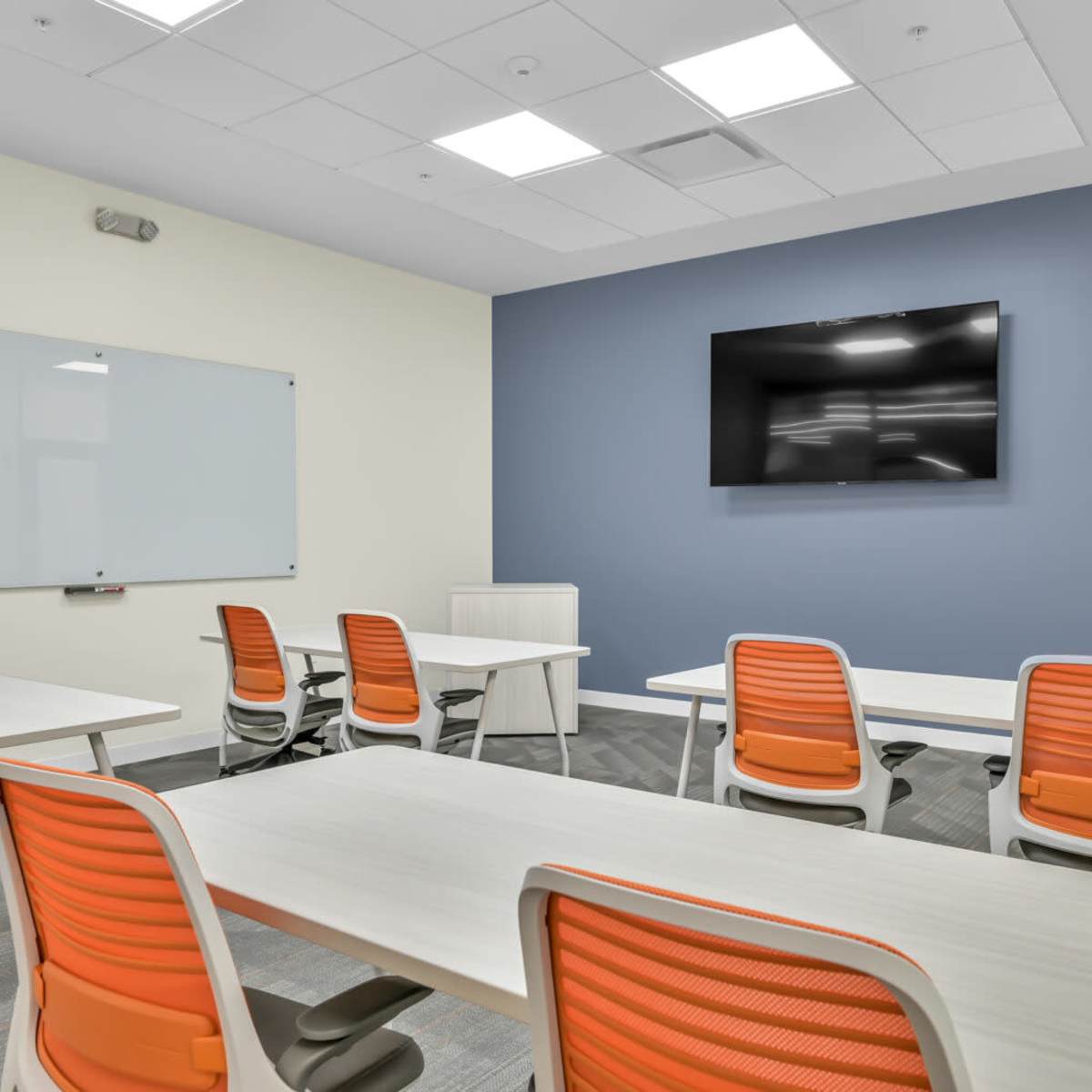 A modern conference room features several tables and chairs with orange accents, a whiteboard, and a wall-mounted television.