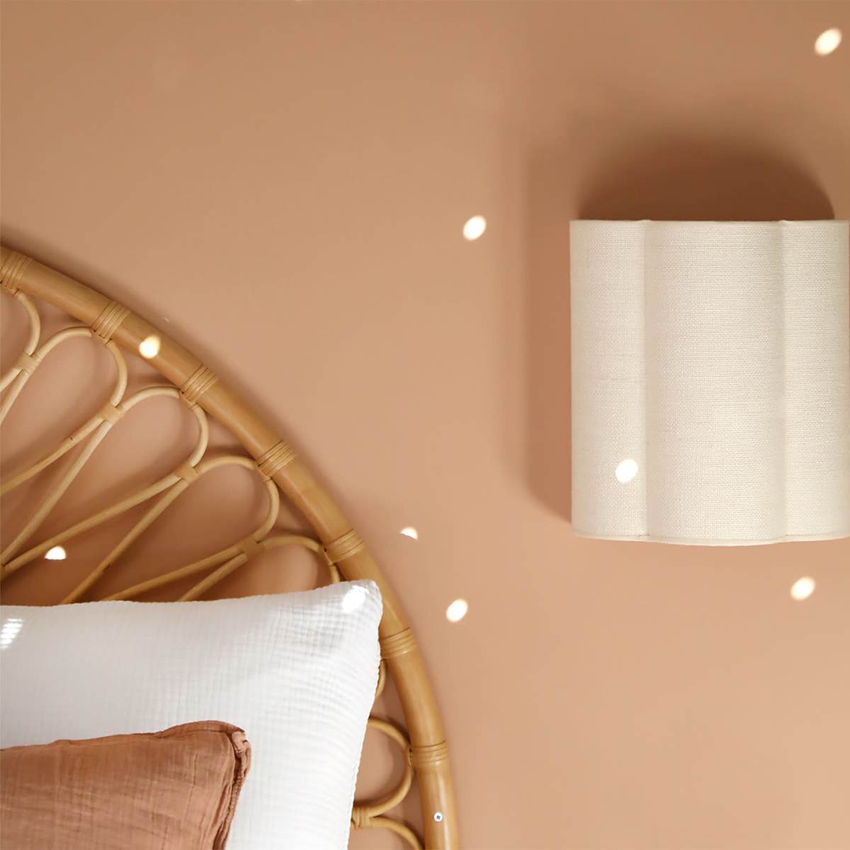A soft, beige wall lamp is mounted on a light brown wall beside a rattan bed headboard with white bedding.