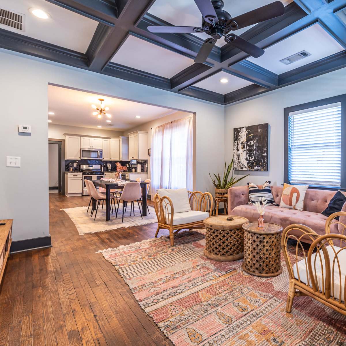 The living room features a pink sofa and rattan chairs, with a large painting on the wall and a view into a kitchen area.