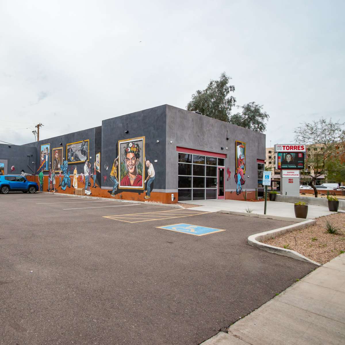 A gray building with large murals on its exterior and a parking lot in front, located on a city street.