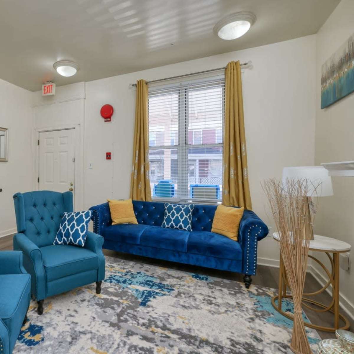 The living room features a blue sofa and two matching chairs, with a patterned rug on a light wood floor and large windows with yellow curtains.