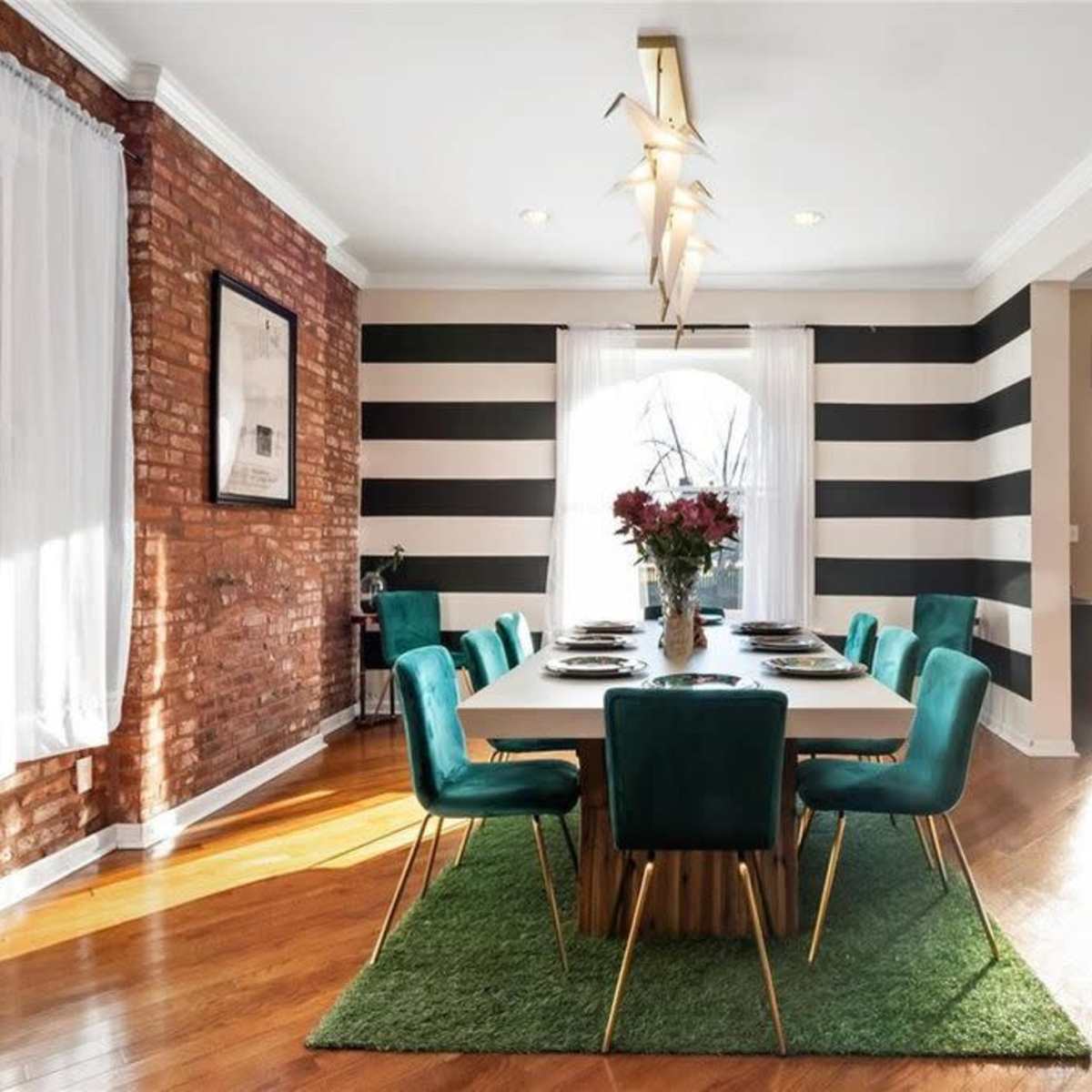 A modern dining room features a large table surrounded by green chairs, with a decorative rug, brick wall, and striped wallpaper.