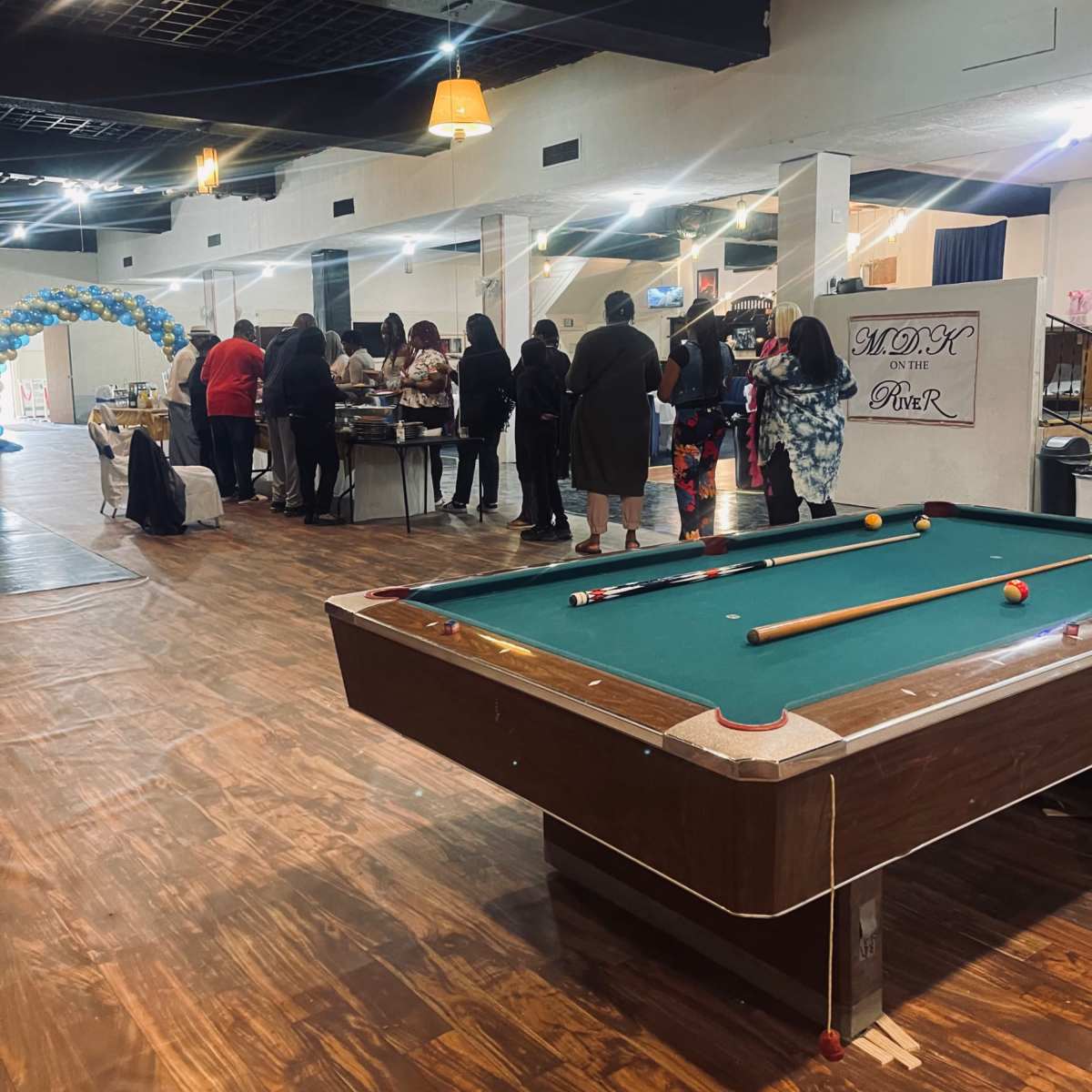 A large indoor space with a pool table in the foreground and a group of people gathered around tables in the background.
