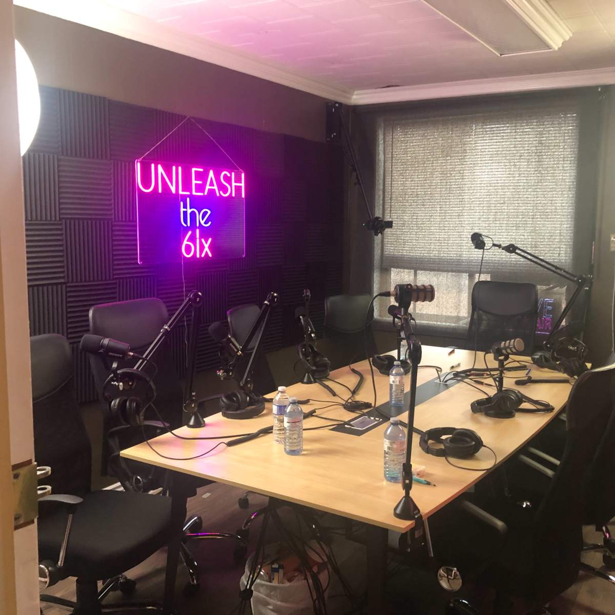 A podcast recording studio featuring a large table surrounded by several chairs, microphones, and a neon sign that reads "UNLEASH the 6ix."