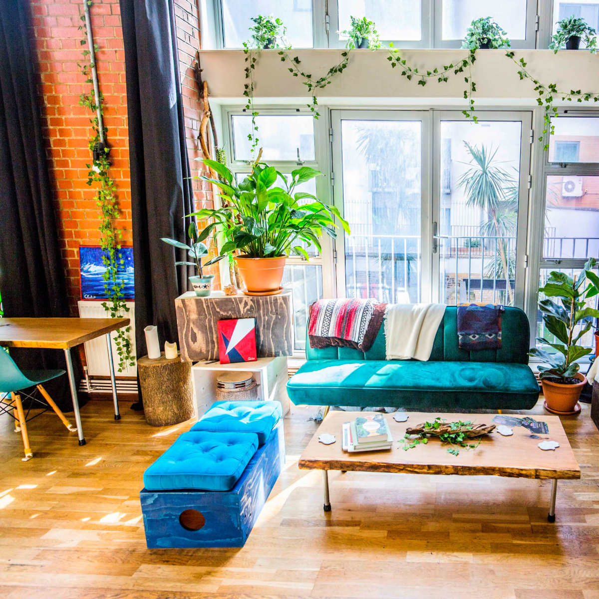 A bright living room filled with plants, featuring a teal couch, wooden coffee table, and large windows allowing natural light.