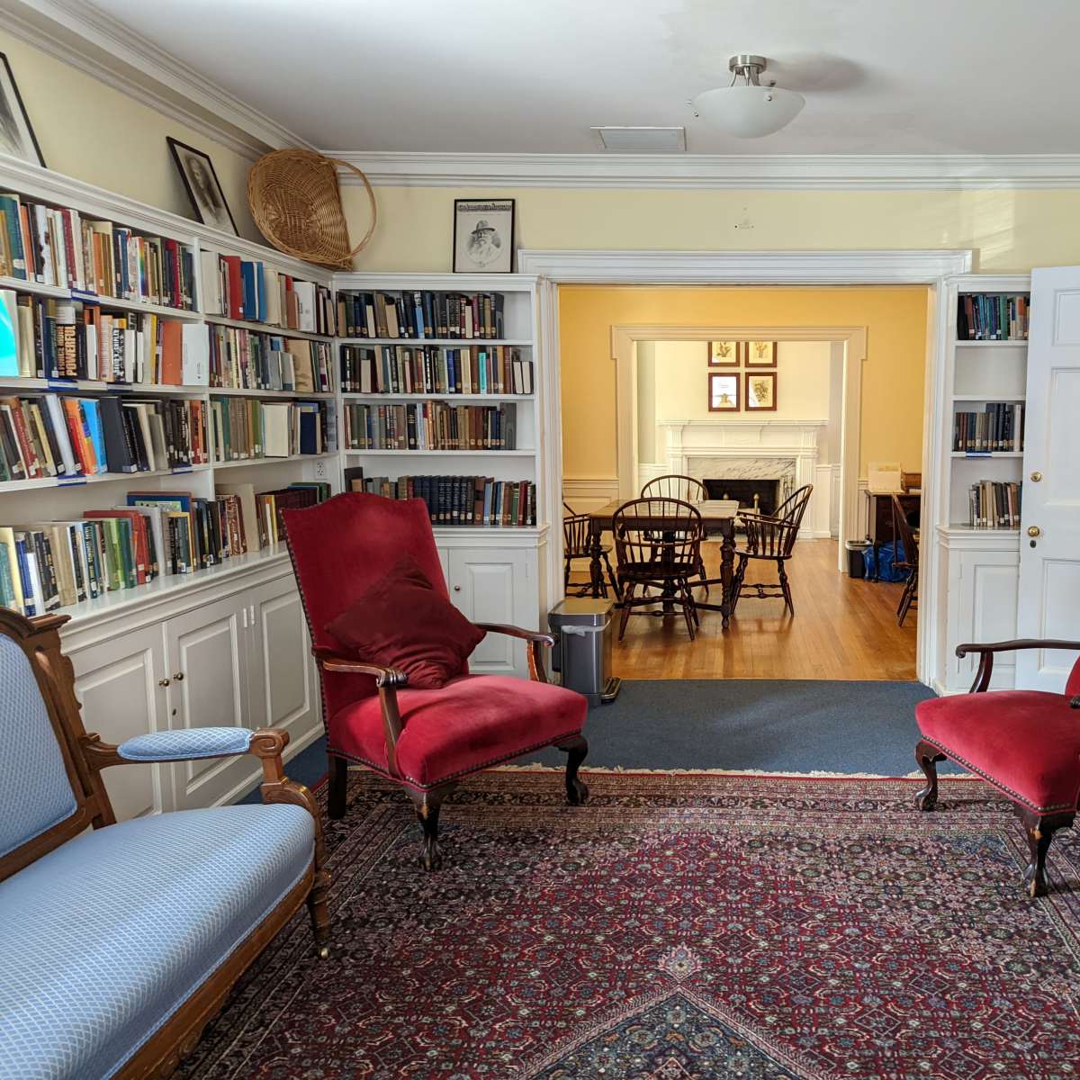 A room furnished with red upholstered chairs, a patterned rug, and bookshelves filled with books lining the walls, leading to a dining area through an open doorway.