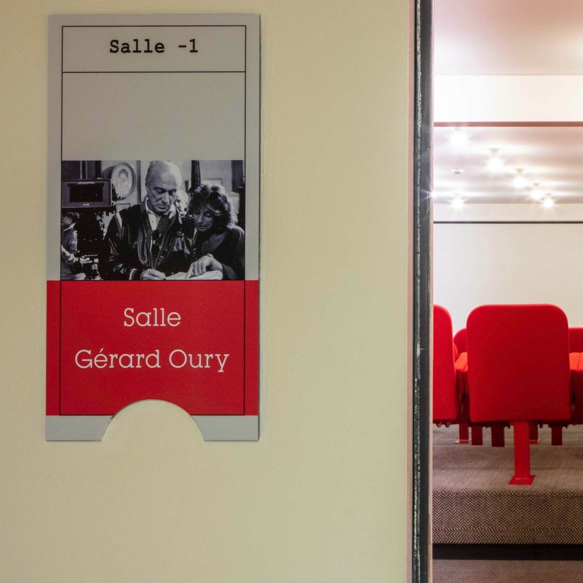 A sign labeled "Salle Gérard Oury" is positioned next to a small theater room featuring red seating and a projector screen.