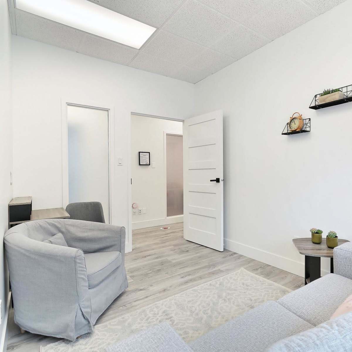 A small, modern waiting room with a gray sofa, a floor lamp, and a table with decorative plants against a white wall.