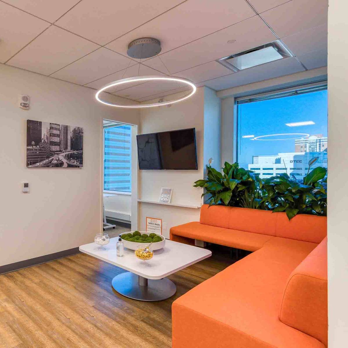 A modern office hallway with orange seating, a wall-mounted TV, and large windows providing natural light.